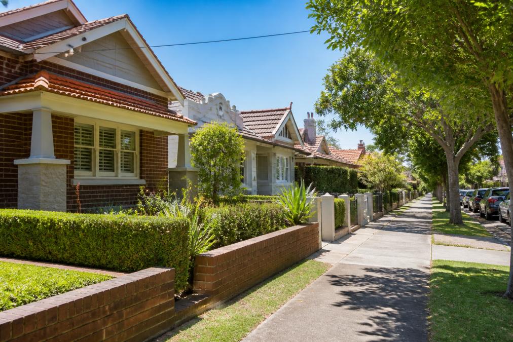 A tree-lined Melbourne suburban street with a mix of period homes and modern renovations on a sunny day, a for-sale sign outside a well-presented brick home with a manicured front garden