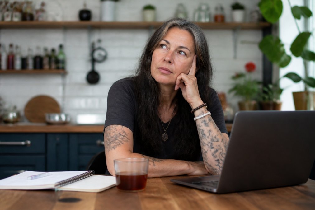 A woman in her mid-40s sitting at a kitchen table with a laptop open, staring into the middle distance with a slightly frustrated expression, a cold cup of tea beside her and a notebook with scribbled reminders nearby
