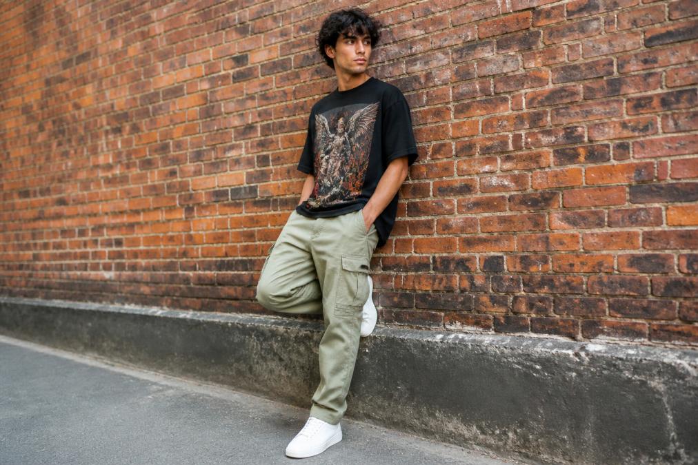 A young man wearing relaxed fit tapered cargo pants, clean low-top sneakers, and a graphic crew-neck tee, standing confidently against an urban brick wall in natural daylight