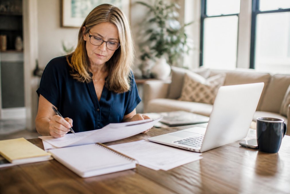 Woman reviewing medical documents and taking notes at home with a laptop open, researching healthcare options in a comfortable, well-lit space