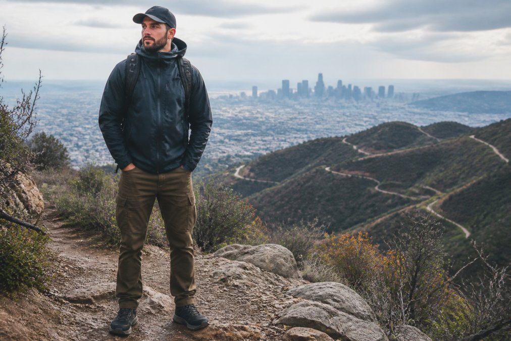 A man in a technical shell jacket and rugged pants standing on a rocky trail with a city skyline visible in the distance, embodying the urban-to-trail transition