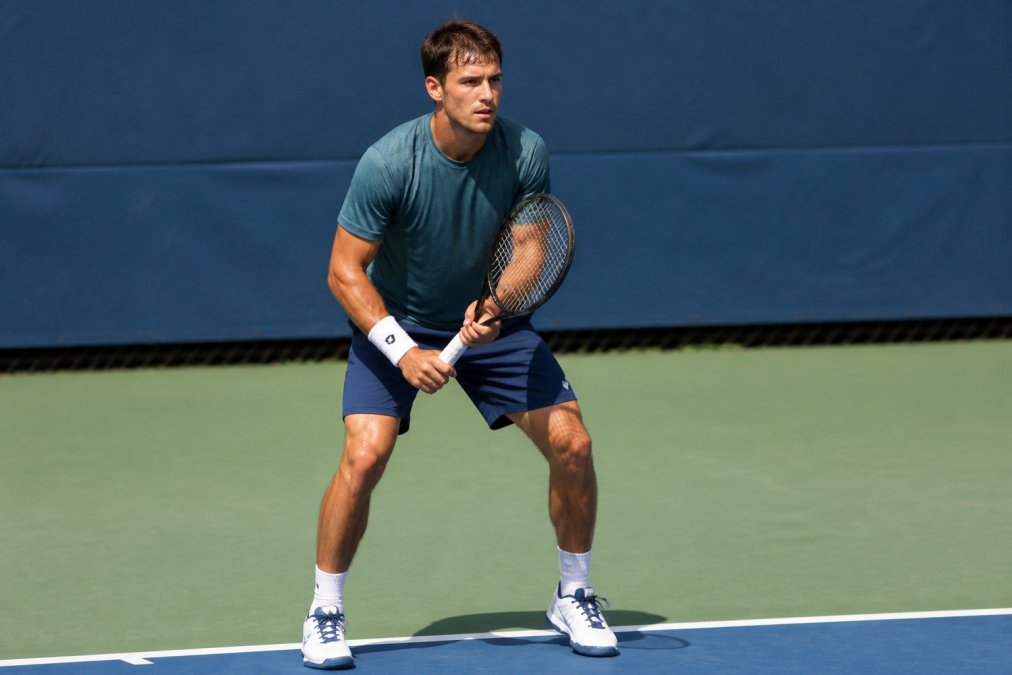 Competitive tennis player at the baseline between points, hand on racket, focused expression and controlled posture, preparing for the next serve in a tiebreaker situation under bright court lighting