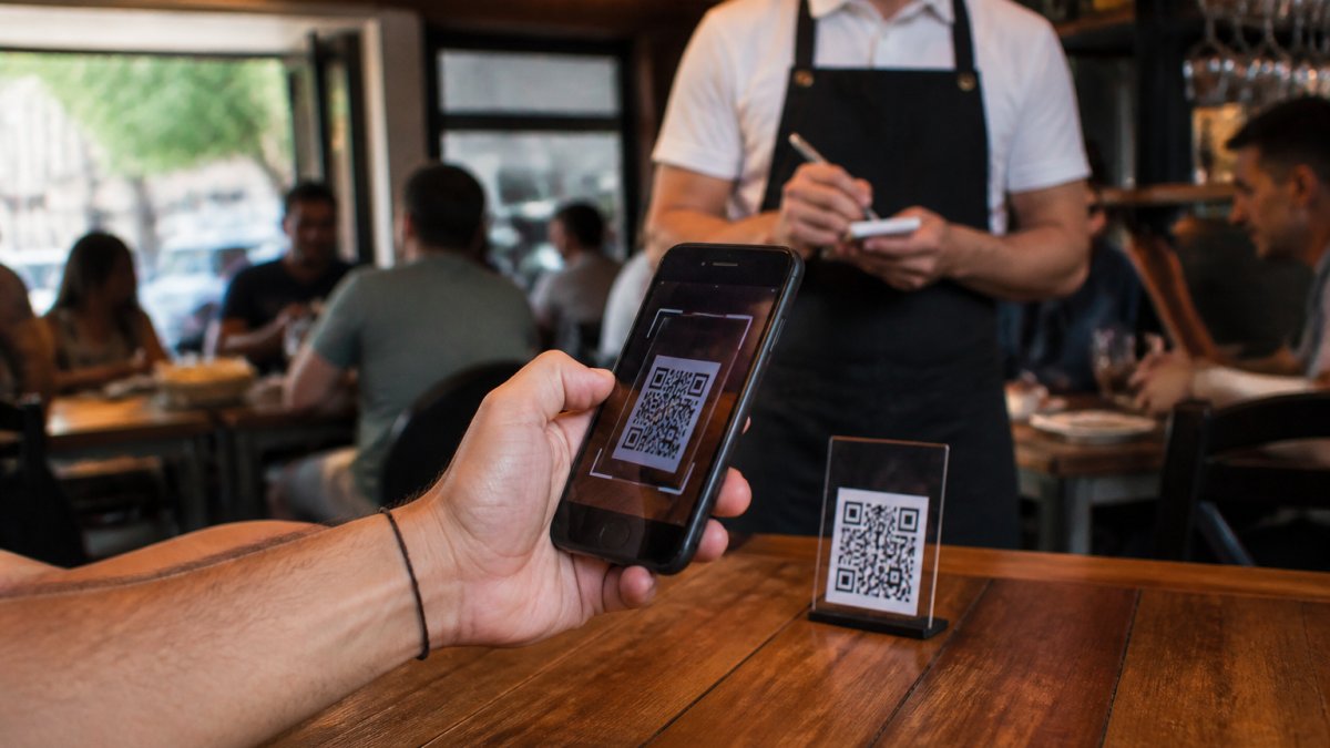 Close-up of a customer's hand holding a smartphone scanning a QR code on a wooden table in a busy restaurant, with a waiter in the background
