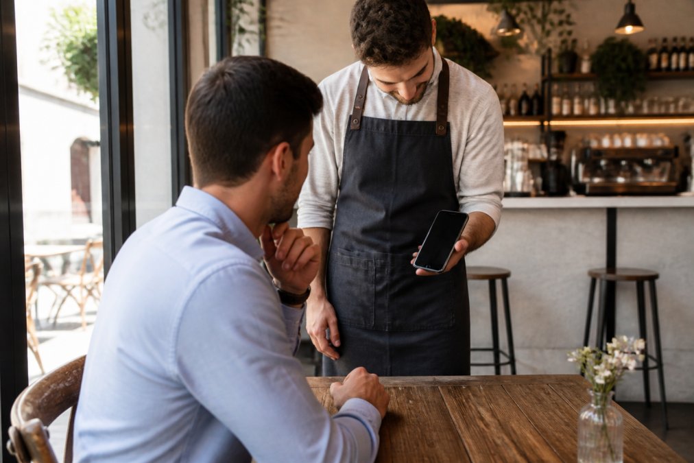 Camarero en una cafetería moderna mostrando un menú digital profesional en la pantalla de un smartphone a un cliente sentado en una mesa de madera, con luz natural entrando por la ventana