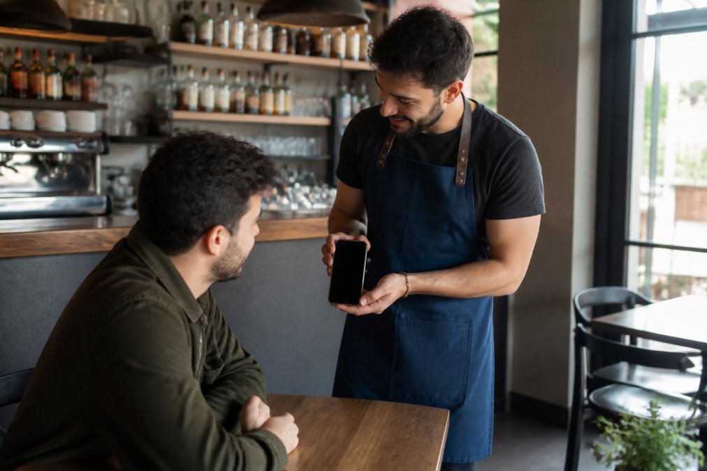 Camarero mostrando un menú digital para cafeterías y bares en un smartphone a un cliente en una cafetería moderna.