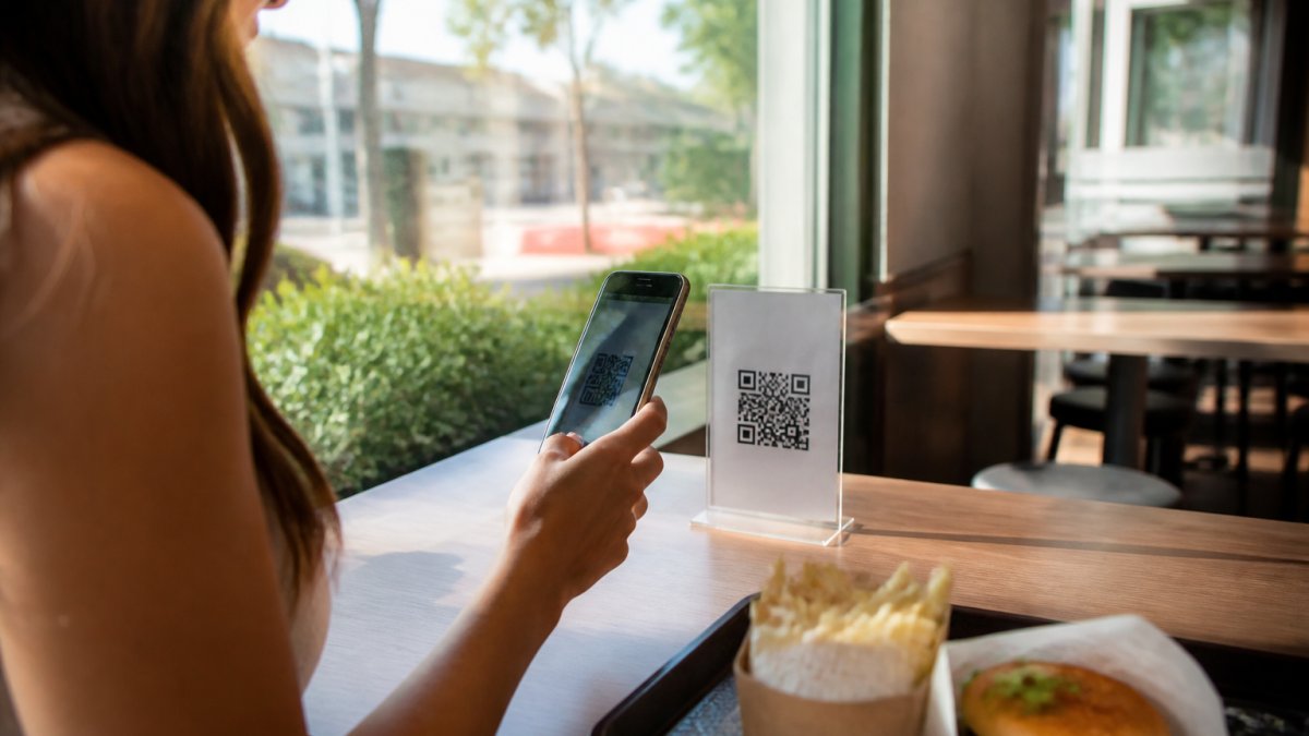 Cliente en un restaurante de comida rápida escaneando código QR en la mesa con su smartphone para acceder al menú digital, luz natural entrando por ventanas, ambiente moderno