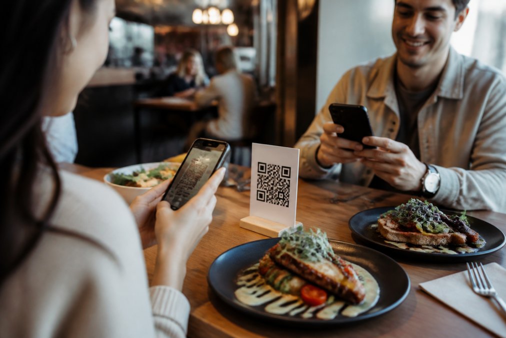 Comensales en un restaurante moderno escaneando un código QR en la mesa con sus smartphones, platos de comida artesanal en primer plano con iluminación cálida