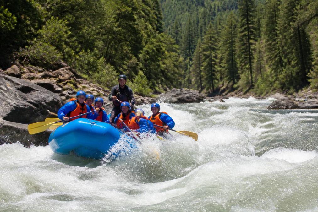 Guided raft navigating Class III rapids during a river rafting trip through a sunny, pine-covered canyon.