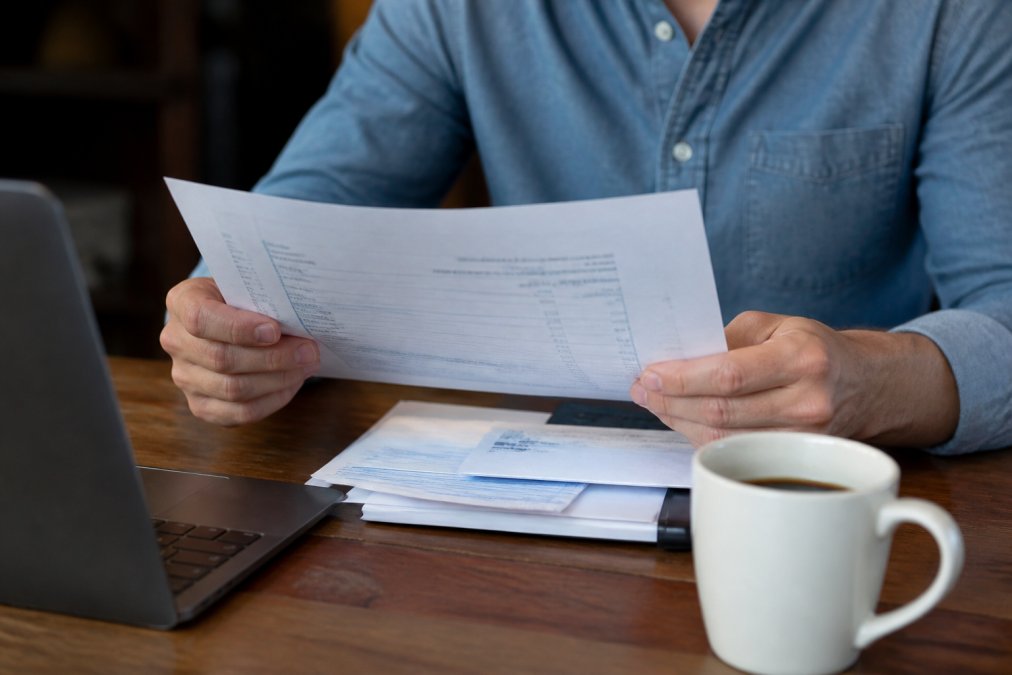 Close-up of a business owner's hands holding a printed merchant statement while reviewing it next to a laptop and a cup of coffee on a wooden desk