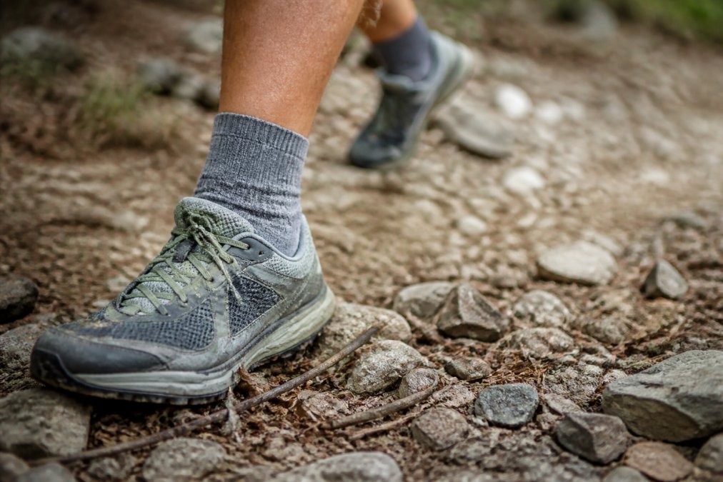 Close-up of a runner's foot in a merino trail running sock during an outdoor run on rocky, debris-filled terrain, showing the sock's protective barrier and fit in action