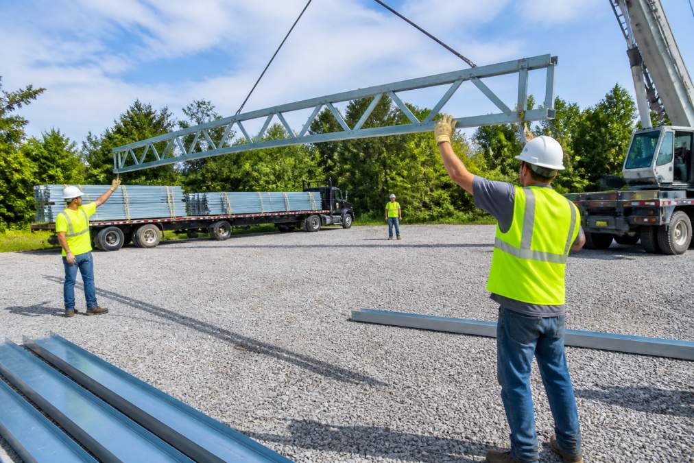 A professional installation crew in hard hats and safety vests guiding a steel truss into place with a crane on a freshly prepared gravel building pad, with a delivery truck and stacked steel panels visible in the background