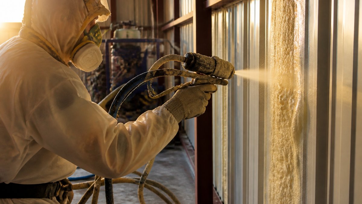 Professional spray foam applicator in protective gear with respirator and chemical-resistant suit applying closed-cell foam to the interior metal wall of a steel building, with foam equipment visible in background