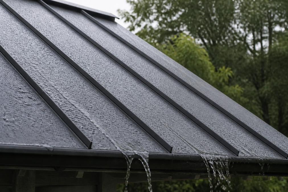 Close-up of a metal roof panel with standing seams during light rain, showing water beading and flowing off the surface with trees visible in soft focus
