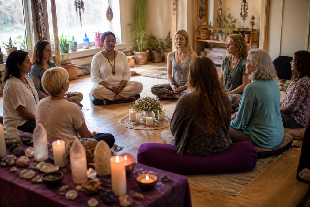 A diverse group of people sitting in a circle during a meditation or spiritual workshop, with crystals and candles on a table nearby, creating a welcoming sacred space with soft natural lighting