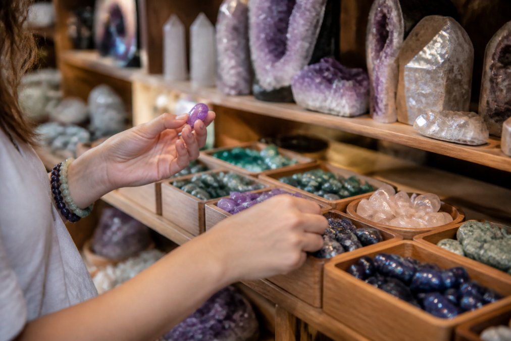 Close-up of hands examining and selecting various polished crystals and gemstones displayed on wooden shelves in a warm, naturally lit metaphysical shop