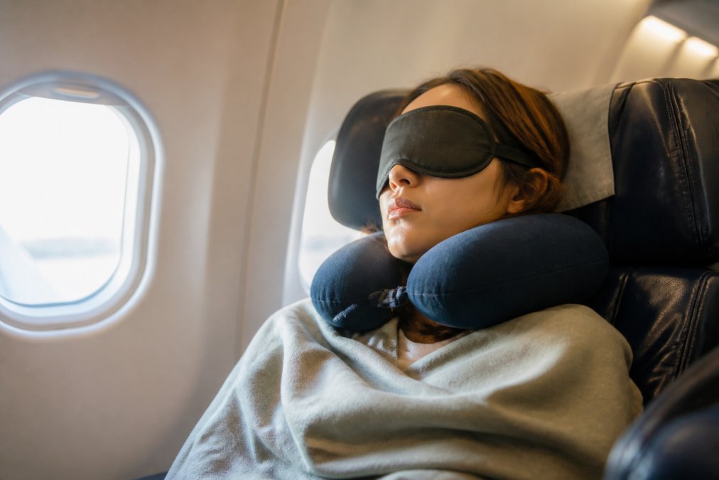 Passenger in an airplane seat wearing a sleep mask and neck pillow, reclined with eyes closed, appearing calm and rested during a long-haul flight