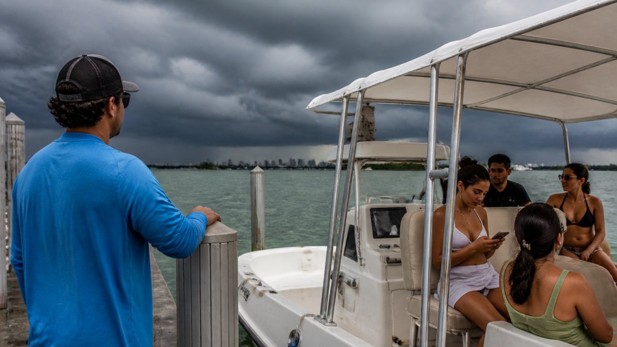 A boat captain on the dock checking dark storm clouds over the water while a group of passengers waits under a canopy, one looking at a phone