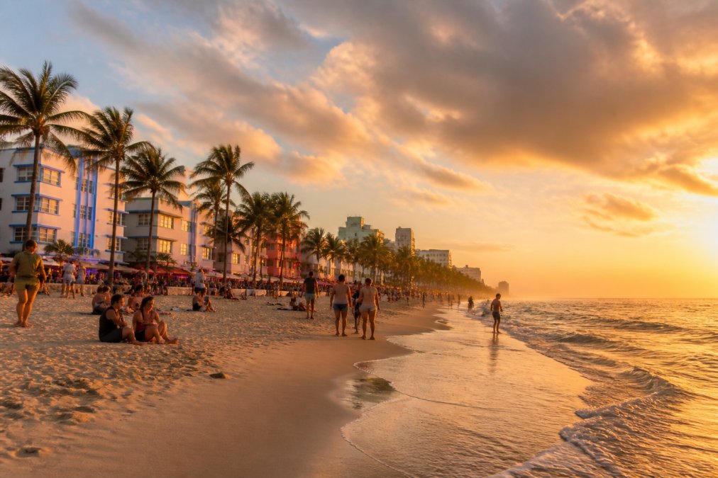 Vibrant Miami beach at sunset with colorful Art Deco buildings lining the waterfront, palm trees swaying, and diverse crowds enjoying the sandy shore with warm golden light reflecting off the ocean