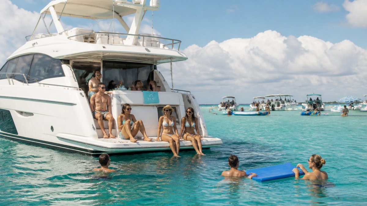 A white motor yacht anchored near a busy Miami sandbar with guests swimming off the swim platform and relaxing on the aft deck under bright afternoon sun