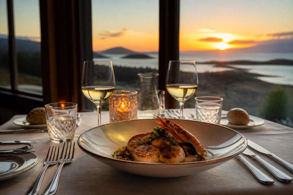 Elegant dining table set with fine glassware and cutlery overlooking a loch view at sunset, candlelit atmosphere with artfully plated fresh seafood and coastal landscape in background