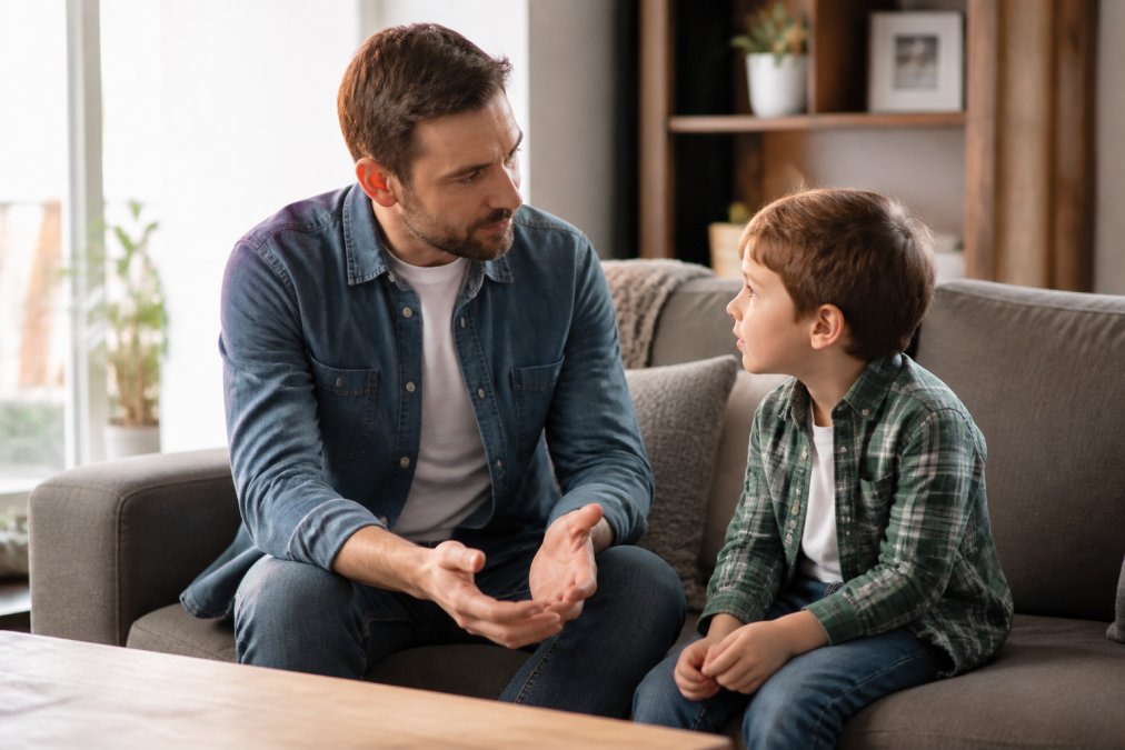 Father and young child sitting together on a couch in a bright living room, having a calm conversation with the child looking engaged and comfortable, suggesting stability and continuity of care during family transition