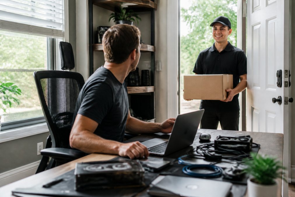 A person working at an organized home office desk with a laptop, multiple tech accessories visible, receiving a delivery box at the door with bright natural lighting from a window