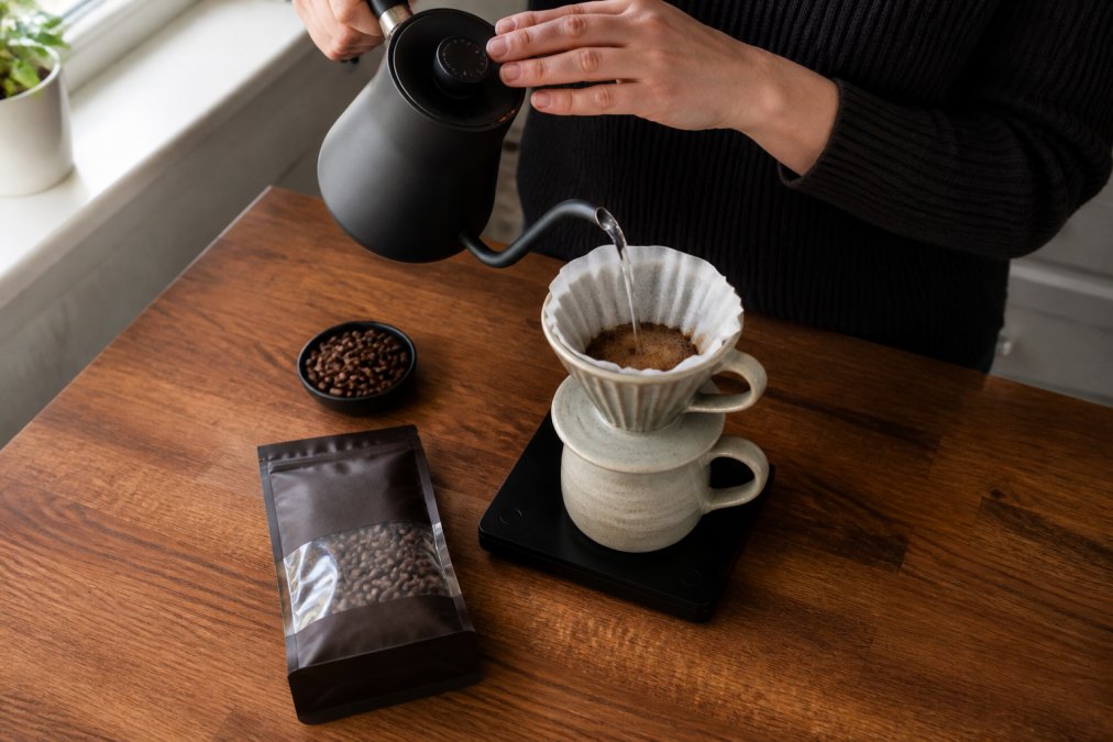 Overhead shot of a person's hands carefully pouring a thin stream of hot water from a gooseneck kettle over a ceramic pour-over dripper, a small sealed bag of specialty coffee beans and a digital scale visible on a warm-toned wooden kitchen counter, soft natural window light