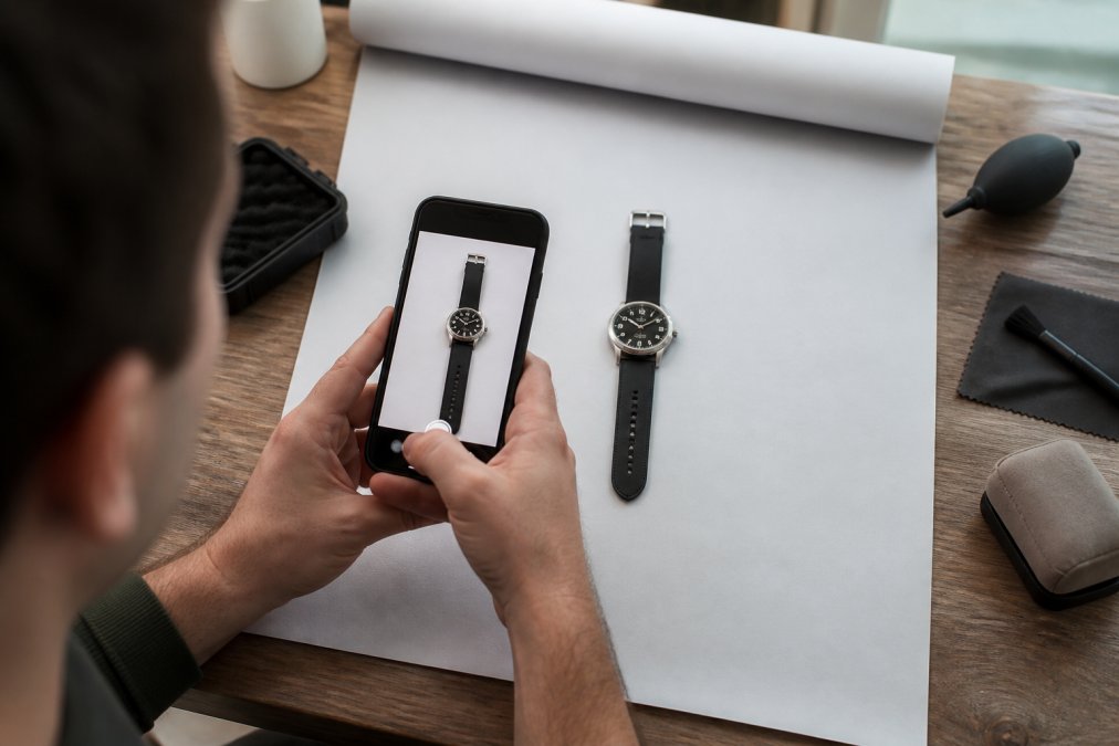 Collector photographing a microbrand watch with a smartphone on a neutral background, positioning the watch to showcase dial detail and case finishing, preparing product photos for online resale listing