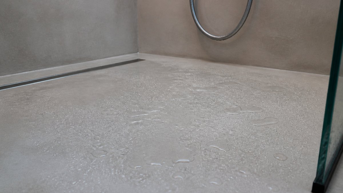 Close-up of a wet bathroom shower floor with water droplets beading on a sealed microcement surface, showing the seamless finish and water behavior on the material in natural bathroom lighting