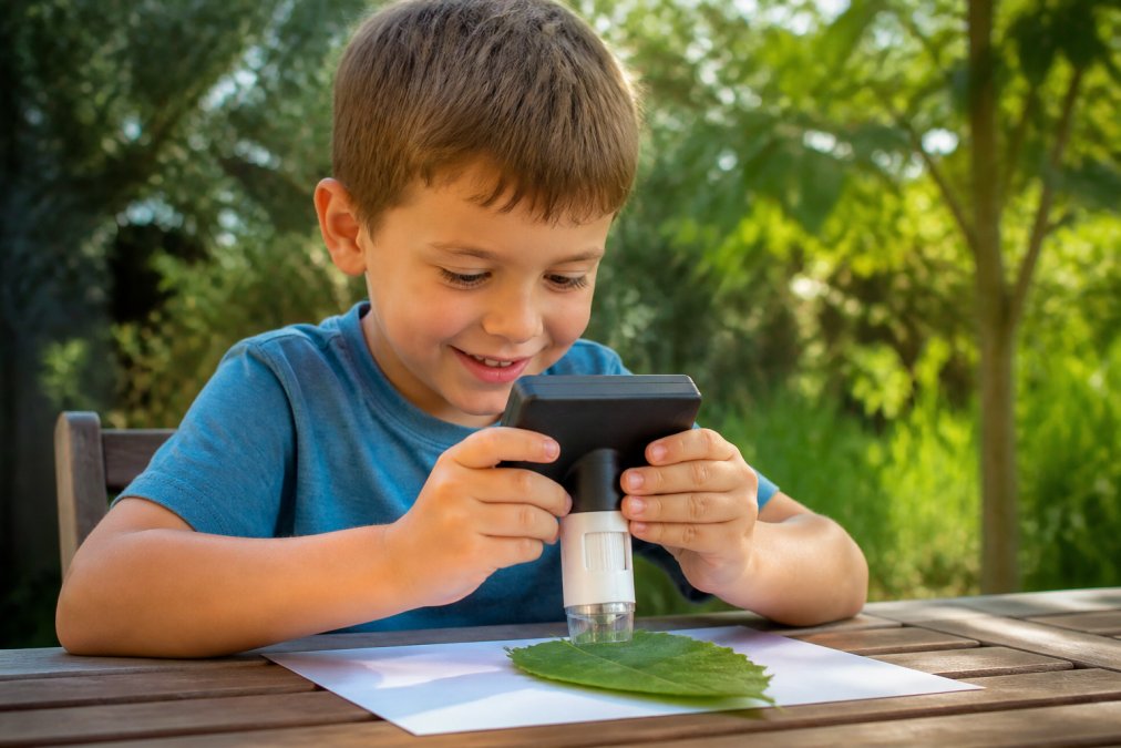 Un enfant d'environ 6 ans, assis à une table en extérieur, tenant un microscope portable à écran avec ses deux mains, observant une feuille posée sur la table, souriant, lumière naturelle du jour