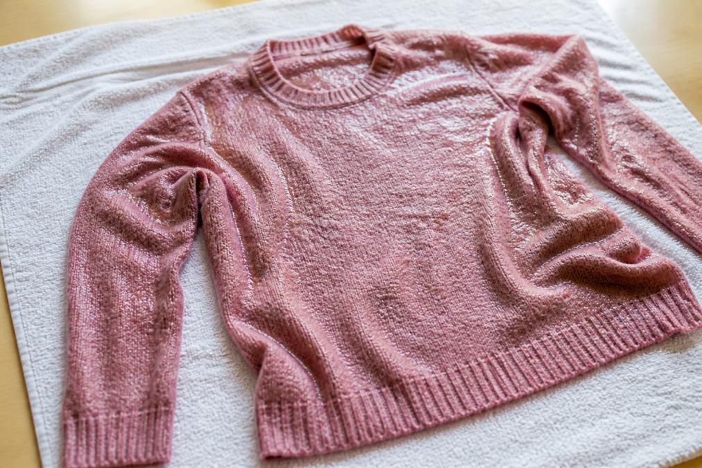 Primo piano di un maglione di lana bagnato appoggiato su un asciugamano bianco disteso in piano, pronto per asciugare in forma, illuminato da luce naturale su una superficie chiara