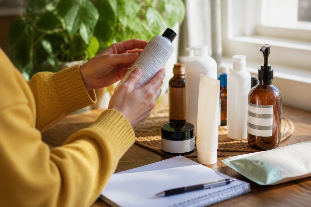 Pet owner's hands holding and carefully reading ingredient labels on multiple pet care product bottles and packages, examining text under natural window lighting while taking notes
