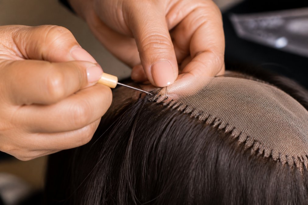 Close-up of skilled hands tying individual strands of human hair to a custom sheitel cap base, showing the detailed knotting technique and realistic hairline construction with natural lighting