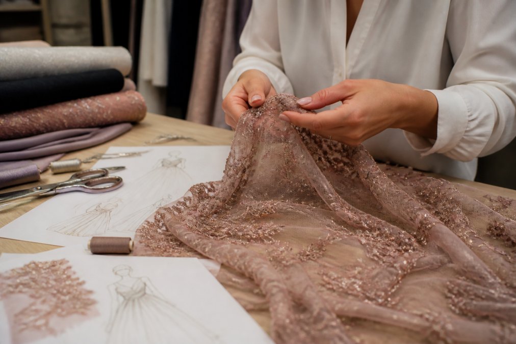 Close-up of a designer's hands feeling the texture and drape of a beaded lace fabric on a worktable, with sewing tools, pattern paper, and sketches nearby in warm studio lighting