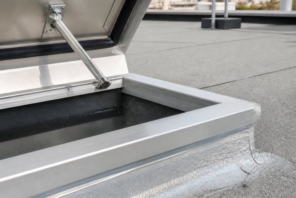 Close-up of roof hatch installed on commercial building rooftop showing weather stripping, galvanized steel frame, and aluminum components with visible corrosion-resistant finish under natural sunlight