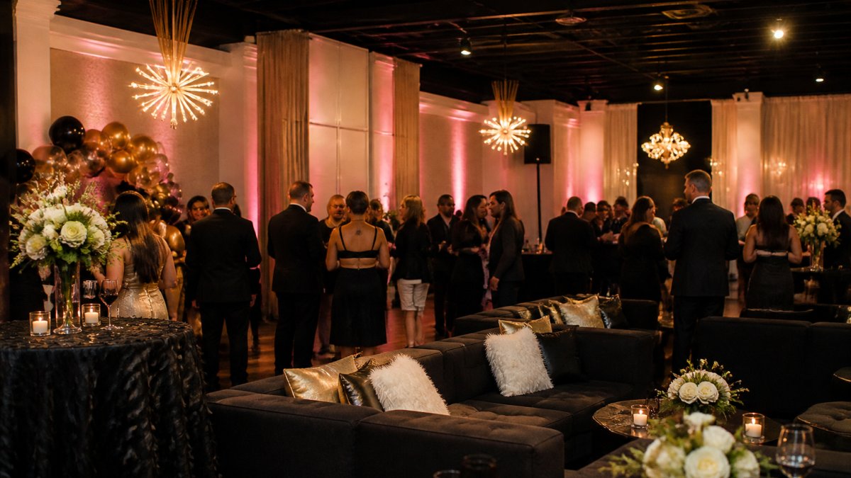 Elegant milestone birthday celebration in progress with guests mingling in a sophisticated lounge setting, featuring professional lounge furniture, ambient up-lighting creating a warm glow, and tastefully decorated tables with centerpieces and overlays