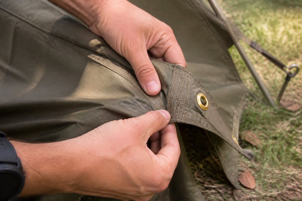 Close-up of hands inspecting a tactical tent's seams, fabric weave, and brass hardware in natural daylight, showing construction details and wear resistance