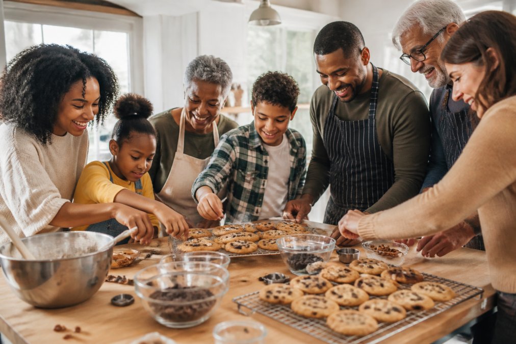 Multi-generational family group of diverse ages and races, including Black family members, gathered around a bright kitchen table with freshly baked cookies cooling on racks, mixing bowls, chocolate chips, and pecans scattered across the counter, natural sunlight streaming through windows