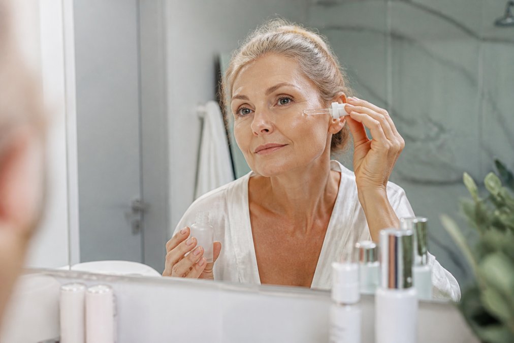 Close-up of a woman in her fifties applying serum to her face in a bright bathroom, soft natural light, focused expression, skincare bottles visible on the counter