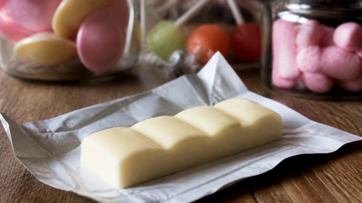 A close-up of an unwrapped Milkybar Choo classic bar resting on a rustic wooden surface, creamy white chocolate texture visible, soft-focus retro sweets in the background under warm natural light