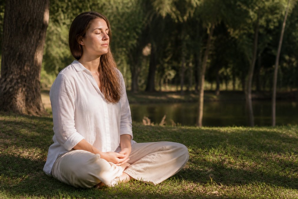 Person sitting outdoors with eyes closed, hands resting peacefully on their lap, practicing mindful breathing in soft natural sunlight with trees in the background