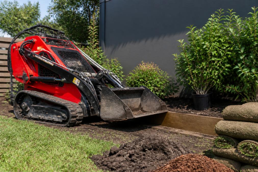 Kanga mini loader positioned in residential backyard with 4-in-1 bucket lowered near garden bed, soil and landscaping materials visible, natural daylight