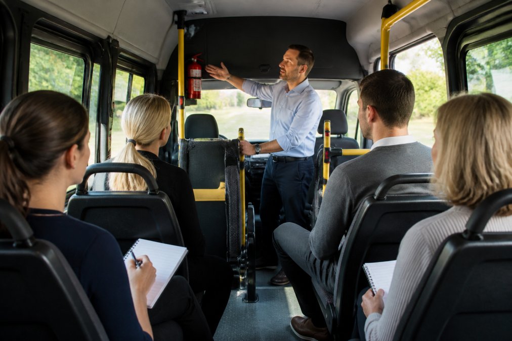 Care home staff members seated in a minibus during a training session, an instructor standing at the front demonstrating the location of emergency exits and safety equipment, natural light streaming through windows, attentive staff taking notes