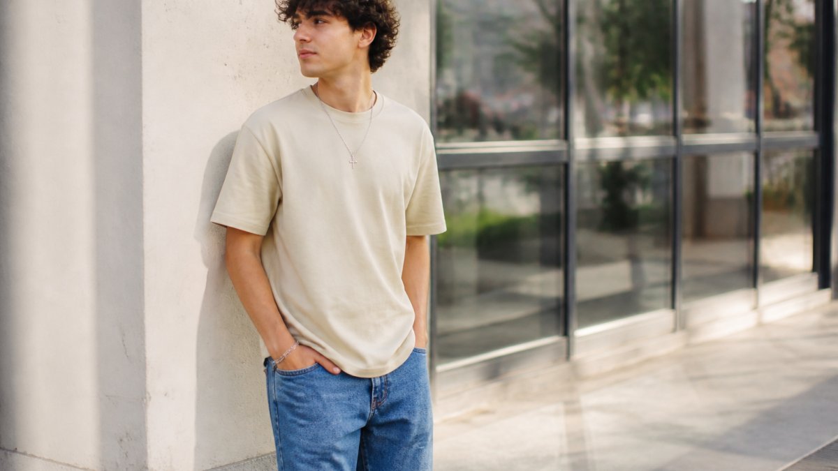 Young man in a neutral graphic tee and jeans standing against a clean urban backdrop in natural light