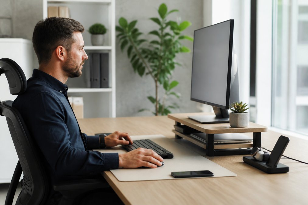Professional working at a clean minimalist desk with elevated monitor stand, wireless peripherals, organized cable management, and a neutral desk mat in a bright modern office