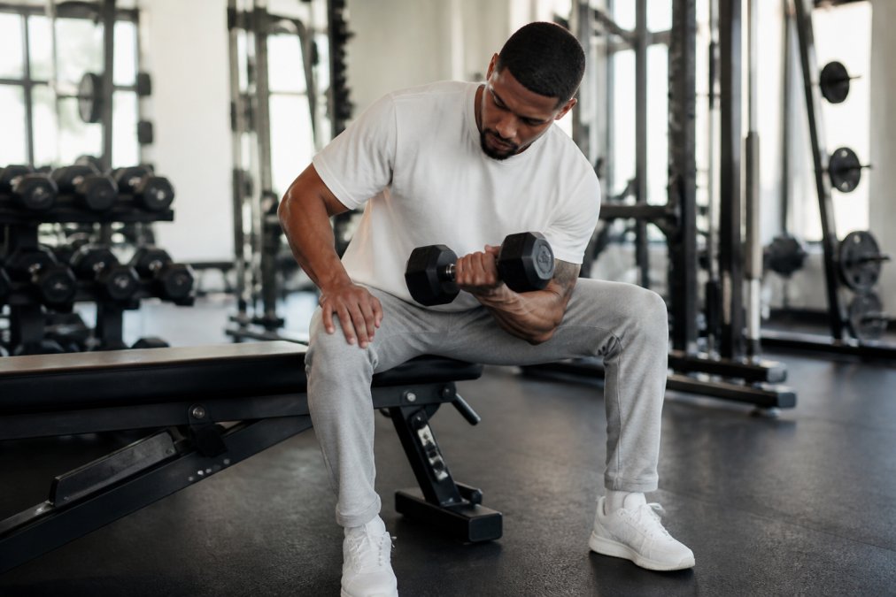 Person wearing a minimalist faith-inspired graphic tee in a gym setting, paired with gray joggers and clean white sneakers, performing a dumbbell exercise with natural gym lighting