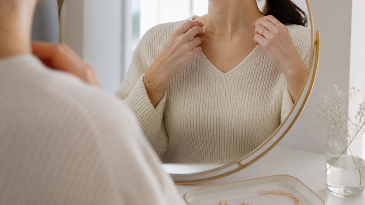 Woman in a cream sweater putting on a thin gold necklace at a sunlit vanity with dainty jewelry pieces nearby
