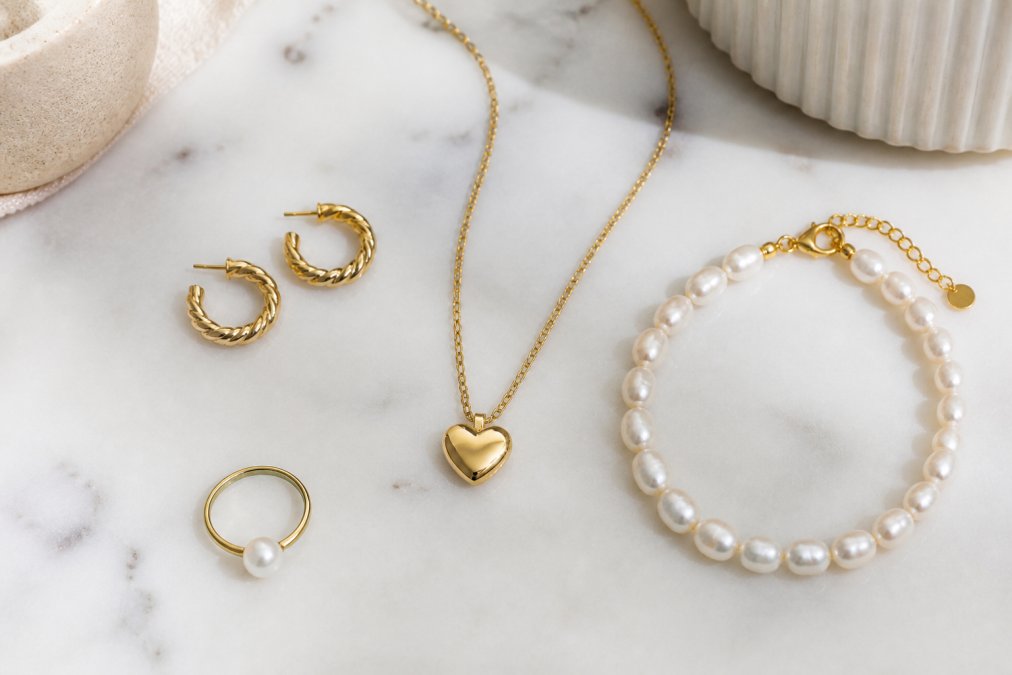 Flat lay of dainty gold jewellery pieces arranged on a white marble surface: small twisted hoop earrings, a delicate heart pendant necklace on a fine chain, a freshwater pearl bracelet, and a simple pearl-topped gold ring, shot in soft natural light