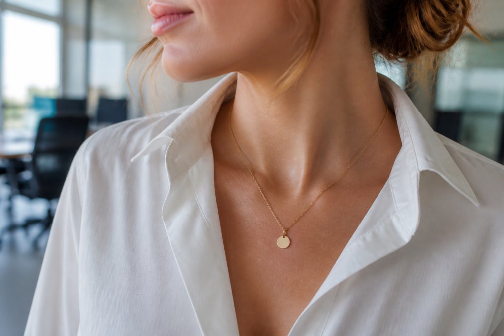 A close-up of a woman's neck and collarbone wearing a delicate 14K gold pendant necklace with a thin chain, styled with a crisp white button-down shirt in a bright office setting