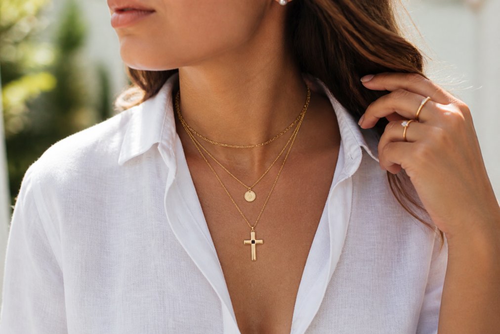 Woman wearing a delicate gold cross necklace layered with other fine chains, photographed in natural daylight with soft focus background showing her wearing a white linen shirt